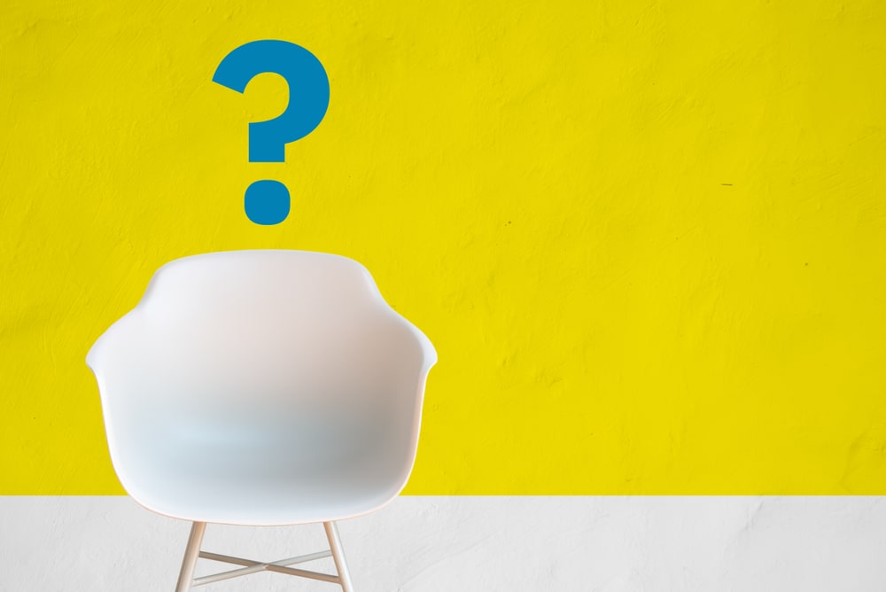 一把白色的现代椅子，靠在黄色的墙上，上面画着一个蓝色的问号。