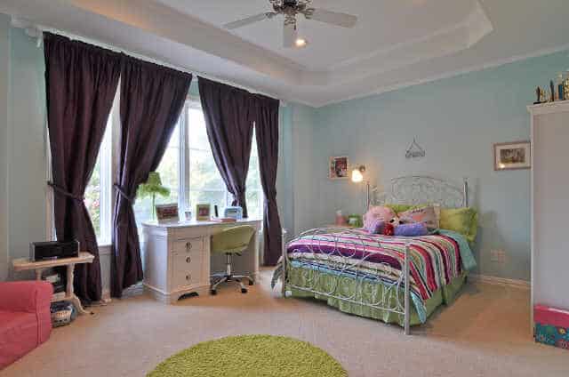 这间卧室有浅蓝色的墙壁、托盘天花板、一张金属床、一张白色的桌子和一张绿色的圆形地毯。