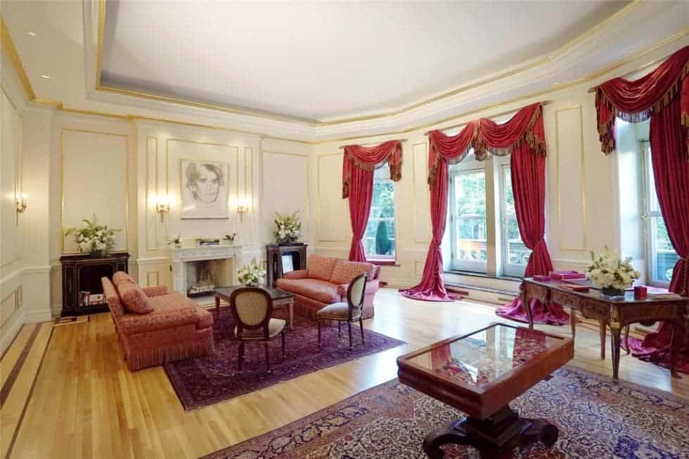 这是一间宽敞的客厅，远处有一座米色的大壁炉，对面是一对沙发，上面有挂着红色窗帘的高窗户。图片来自Toptenrealestatedeals.com。
