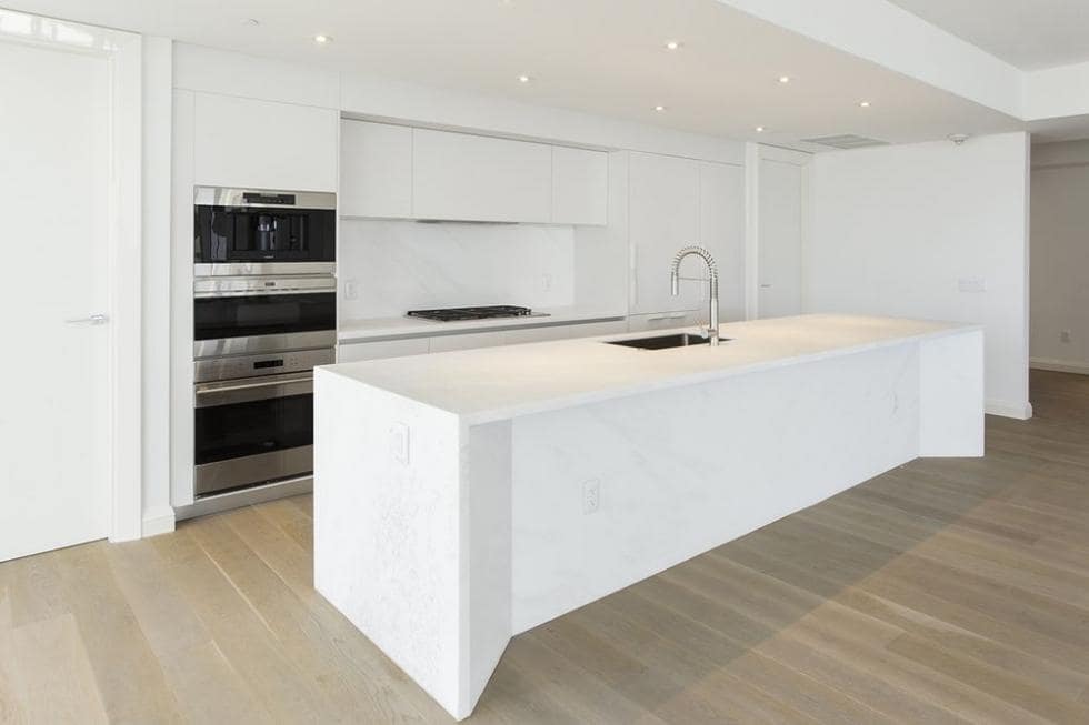 这是一个明亮的白色厨房，有一个大的白色厨房岛，与白色橱柜相匹配，辅以硬木地板和不锈钢用具。图片来自Toptenrealestatedeals.com。