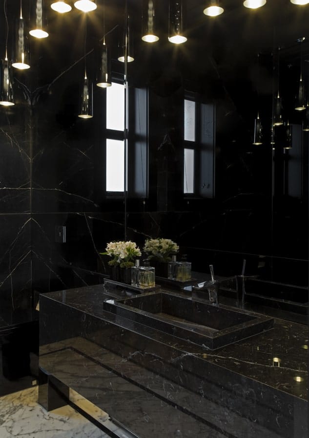 这是房子里浴室的近距离观察，它的梳妆台、墙壁和水槽台面上都有黑色大理石色调。这些都是由吊坠灯的温暖光线补充。