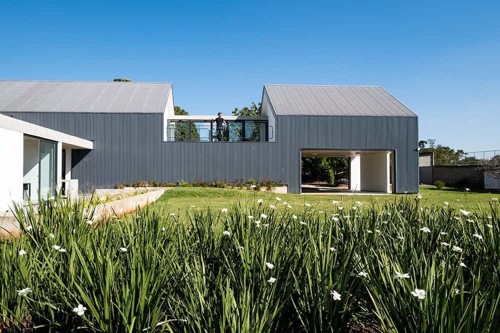 从房子的后面看，它的外观是灰色的，辅以独特的设计和郁郁葱葱的草坪和开花的灌木景观。