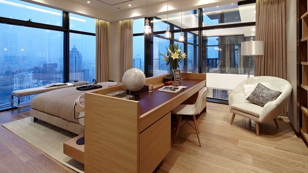 这是一间卧室，床对着远处的大玻璃墙。这是与作为床头板和书桌的大型木结构相连的。
