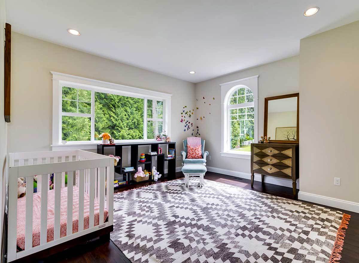 这间卧室有米黄色的墙壁，白色的婴儿床，木制的梳妆台，一张蓝色的躺椅，上面放着一张有图案的大地毯。