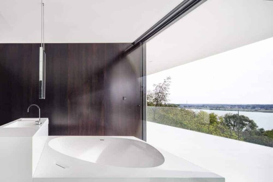 这是一个极简主义浴室的近距离观察，一个巨大的白色结构容纳了浴缸和水槽，与深棕色的墙板形成对比，并被玻璃墙照亮。