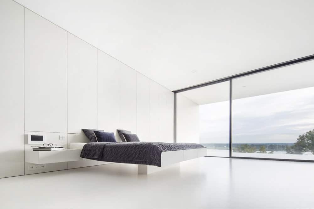 这是一间宽敞明亮的极简主义卧室，白色墙壁始终如一。地板和天花板由现代浮台床的深色床单形成对比。