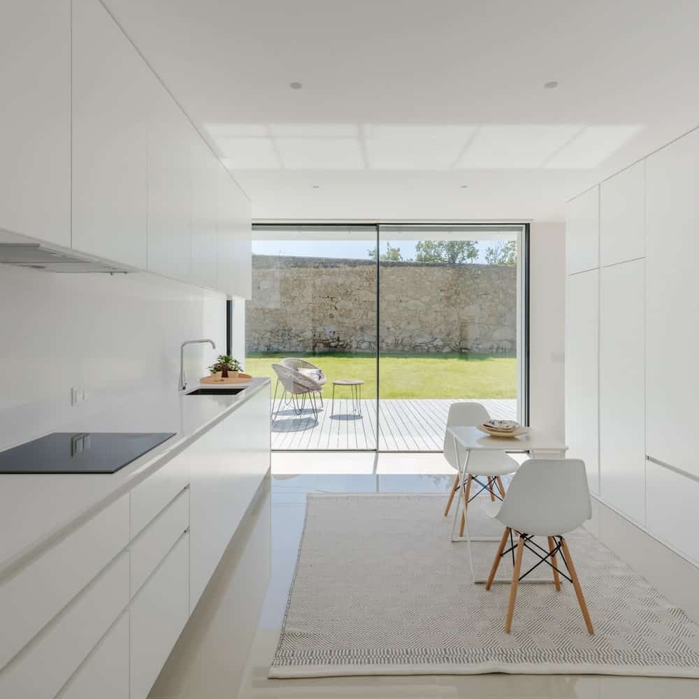 这是一个明亮的白色厨房，在橱柜和早餐角落一致的白色现代色调，玻璃墙壁的自然光照亮了。