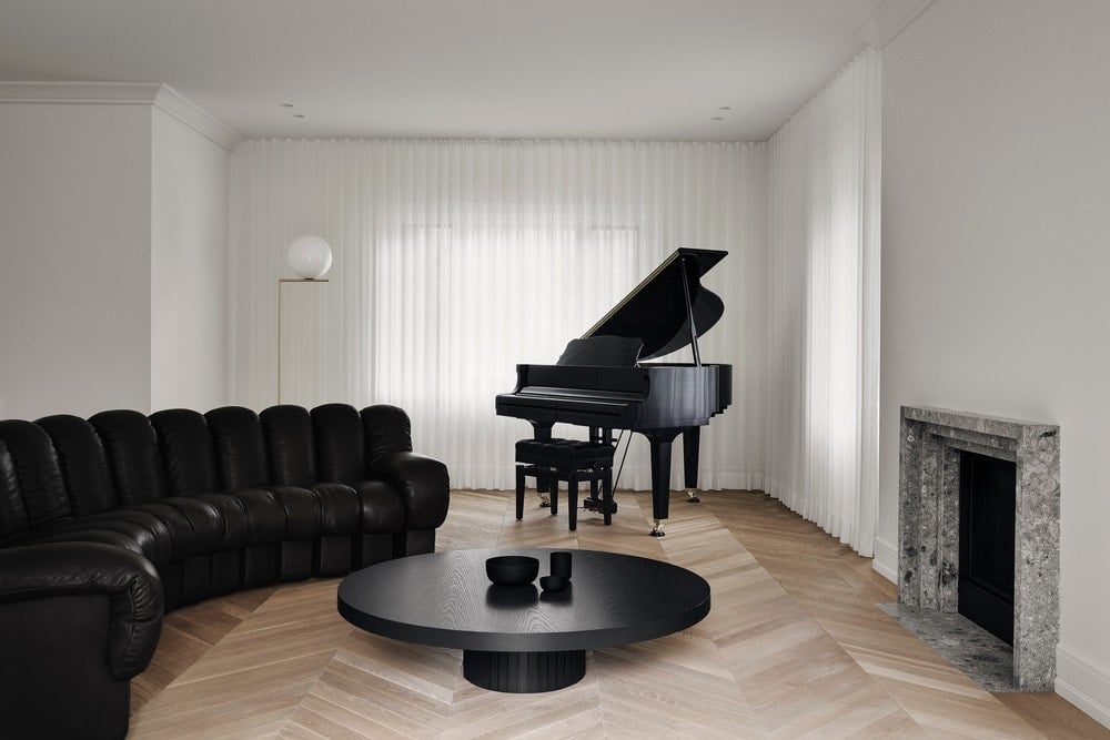 这是一个简单的极简主义客厅，有一个大弯曲的黑色簇绒沙发，搭配黑色圆形咖啡桌和三角钢琴。这些与明亮的白色墙壁和白色天花板形成对比。