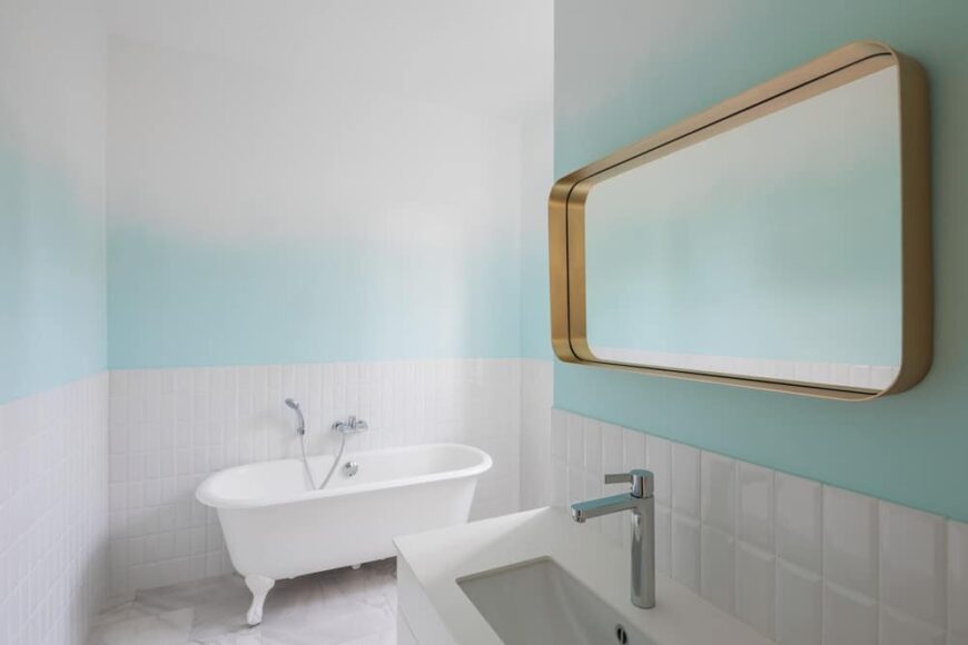 这个极简主义的浴室有一个白色的独立浴缸，一个白色的水槽和白色的瓷砖，与上面墙壁上柔和的色调相辅相成。