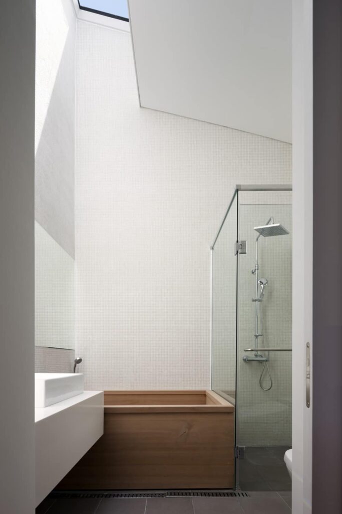 这是主浴室的近景，有高高的拱形天花板、白色墙壁、一个玻璃围成的淋浴区，白色水槽旁还有一个木制浴缸。