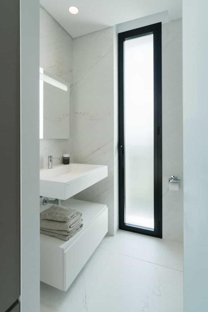 这是一间简单的极简主义浴室，有一个纯白色的浮动水槽和梳妆台，淋浴区磨砂玻璃门侧面有一面无框镜子。