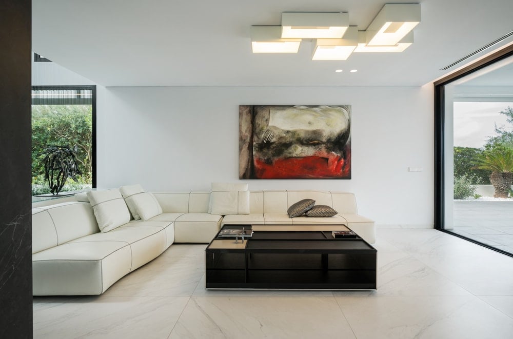 这是一间明亮的极简主义客厅，里面有一张白色的l型组合沙发，与白色的墙壁、天花板和地板相呼应。这些与墙上的艺术品、黑色咖啡桌和上面的现代照明形成对比。