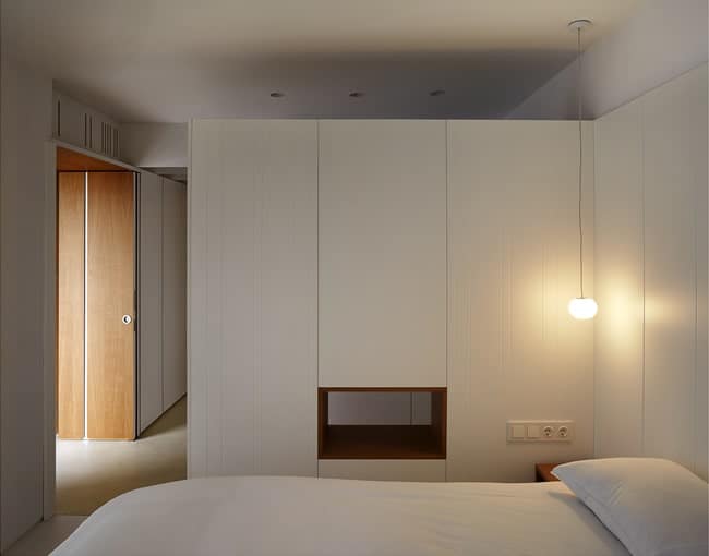 这是一张简单的极简主义床，白色的橱柜和墙壁与白色的床相匹配。床头柜上温暖的灯光作为补充。