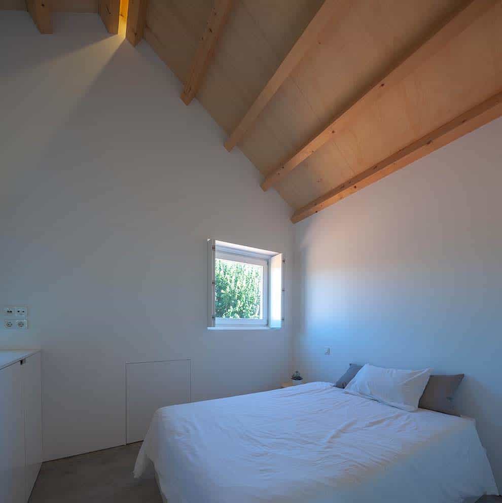 这是一个极简主义的卧室，白色的床与白色的墙壁相匹配，顶部是木制的大教堂天花板，横梁暴露在外。
