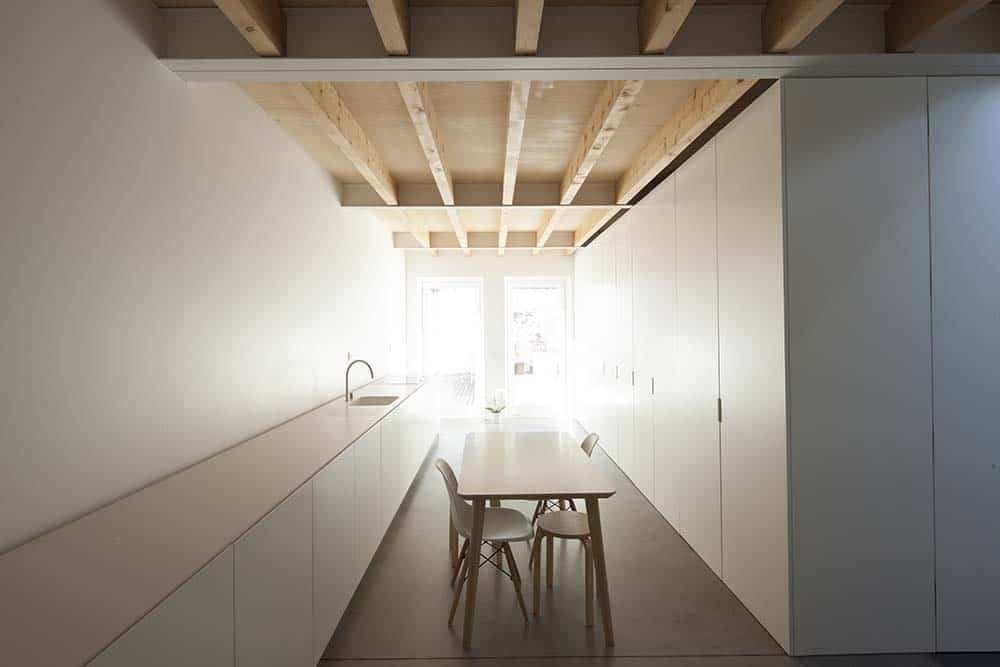 这是一个狭长的可在里面用餐的厨房，两边都是极简主义设计的白色橱柜，木质横梁天花板，中间是一张小桌子。