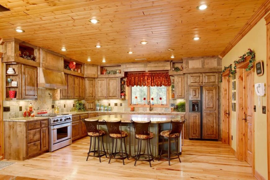 这是一座山上的山屋式厨房厨房，带木制毛绒墙和天花板，搭配木制橱柜衬砌墙壁和厨房岛屿。然后由天花板的多个凹陷灯互补。