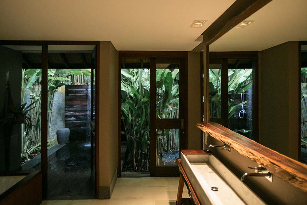 这是浴室水槽的另一个视图，远侧有一扇玻璃门，通向被植物和树木包围的室外淋浴区。