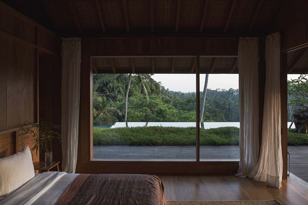 卧室的一侧是一面巨大的玻璃墙，让自然光进入，并提供了周围绿草和热带树木景观的视野。