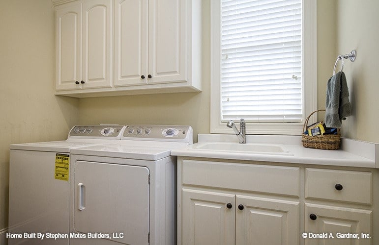 杂物间配有白色洗衣机和烘干机、白色橱柜、瓷水槽和百叶窗。