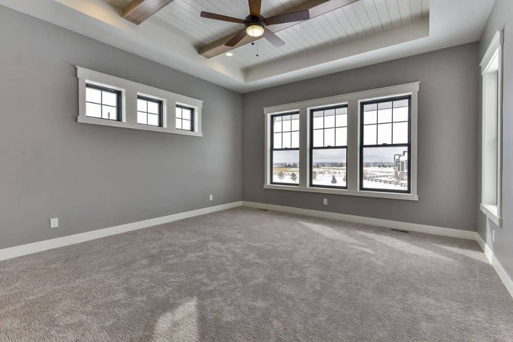 主卧室有灰色墙壁，地毯地板，横梁托盘天花板，和许多框窗。
