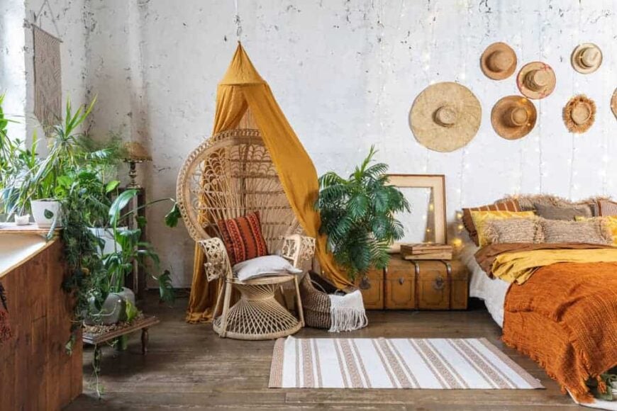 这是一间波西米亚风格的卧室，床上有挂在墙上的帽子，旁边还有一把编织的柳条椅。