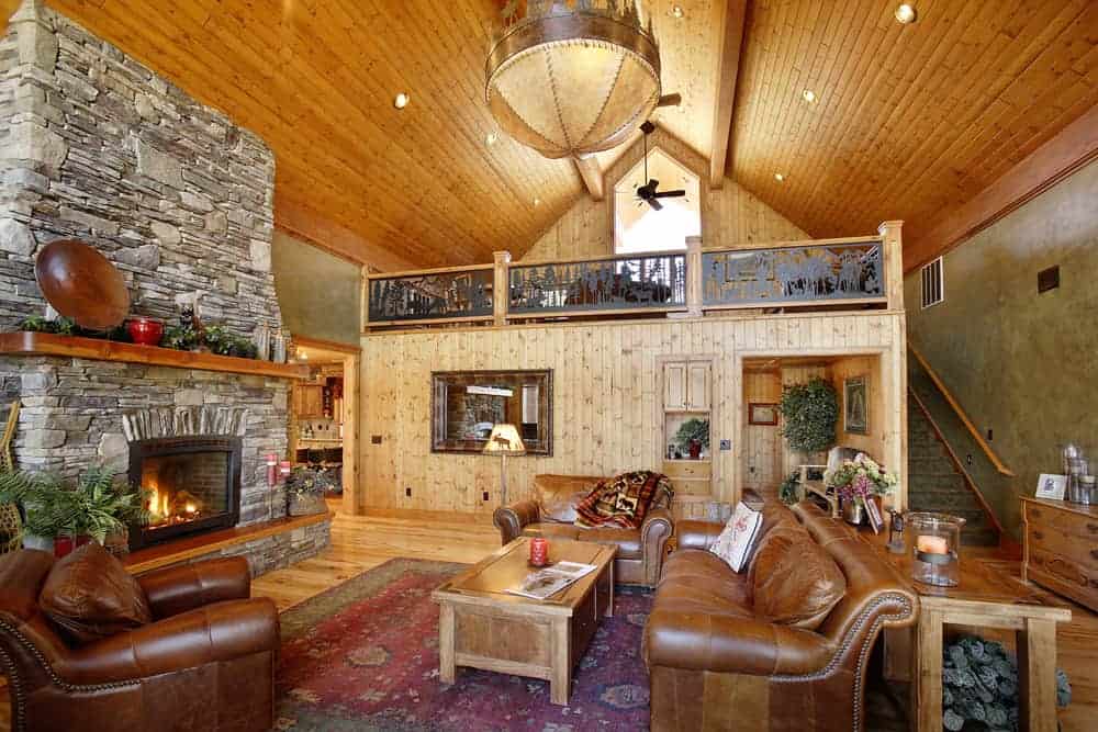 这是一间山间小木屋风格的客厅，有高高的木制大教堂天花板，棕色皮革沙发，木制咖啡桌对面有一个巨大的马赛克石壁炉。