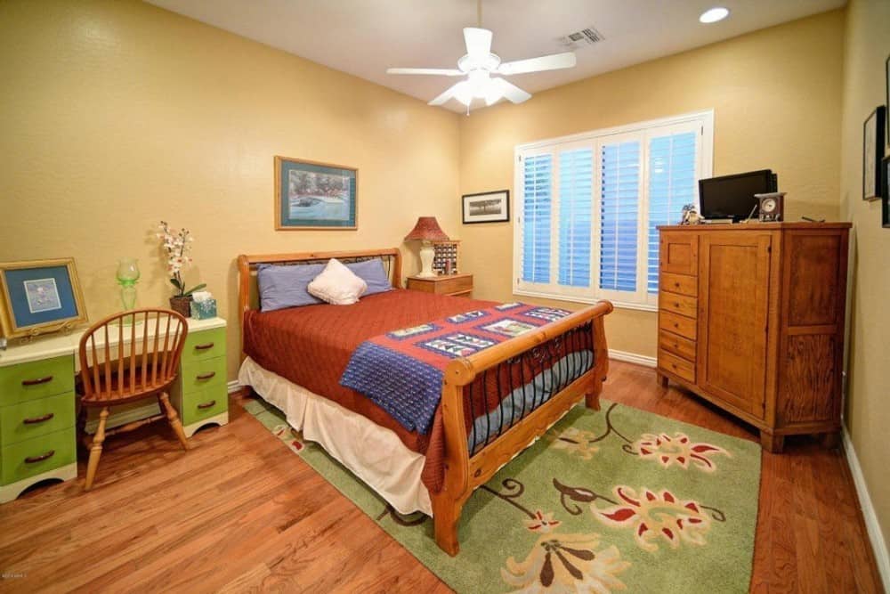 这间卧室有米黄色的墙壁，一张绿色的书桌，一张与硬木地板相匹配的木床，上面铺着碎花地毯。