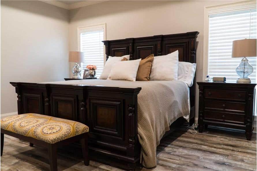 主卧室有一张深色的木床，配套的床头柜，和一个有图案的脚凳。