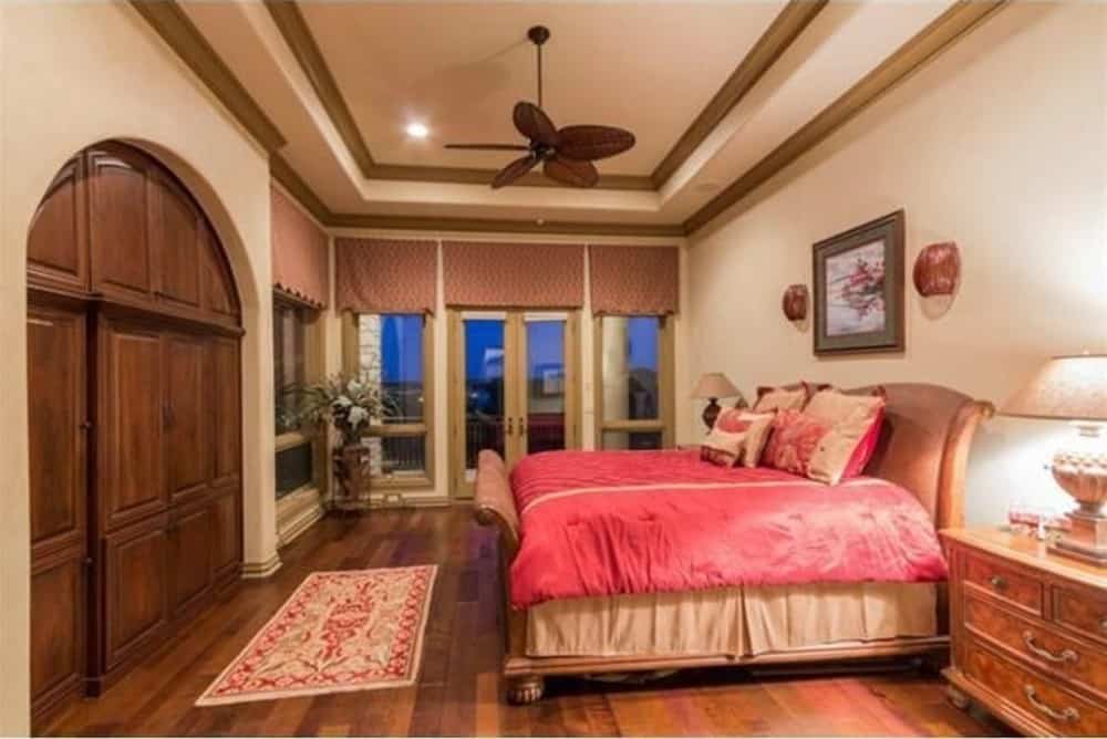 主卧室有一张漂亮的木床，一个红色图案的床架，一个吊扇固定在一个托盘天花板上，天花板上有木饰边。