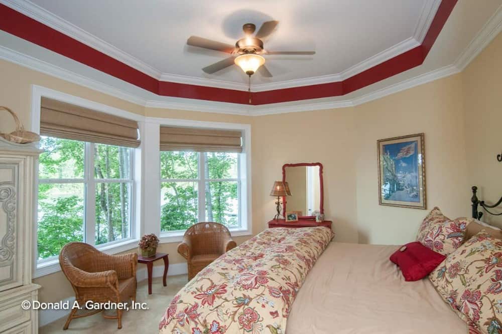 卧室有奶油色的墙壁，托盘天花板，柳条扶手椅搭配一个木制的边桌。